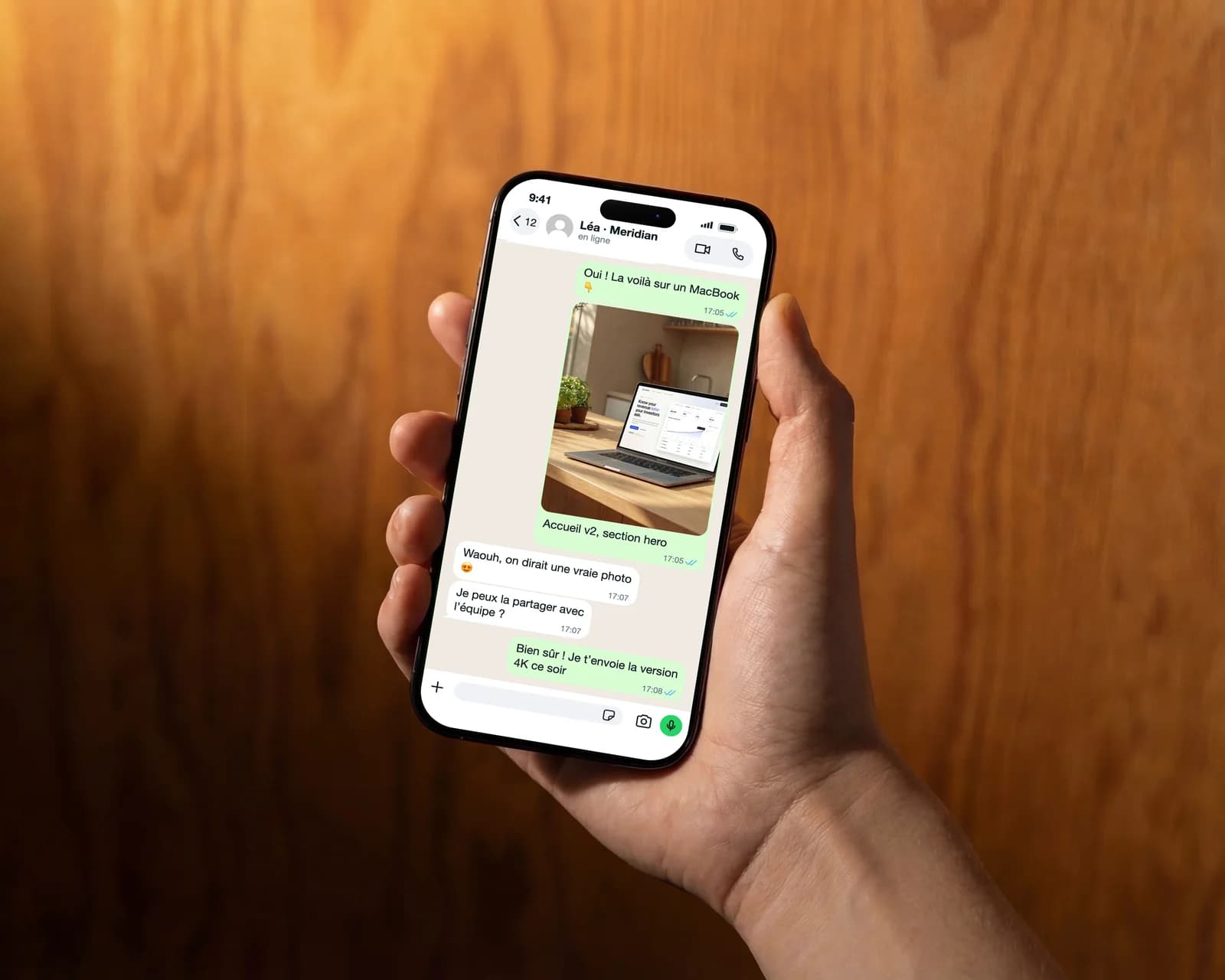 Main tenant un iPhone devant un mur en bois, affichant une conversation WhatsApp où une designer envoie l'aperçu d'une page d'accueil sur un MacBook