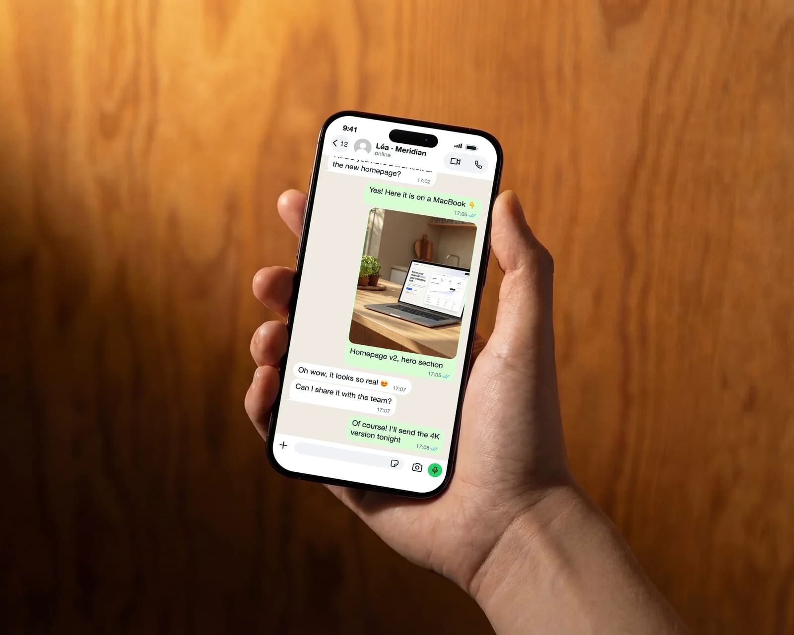 Hand holding an iPhone in front of a wooden wall showing a WhatsApp conversation in which a designer sends a homepage preview on a MacBook