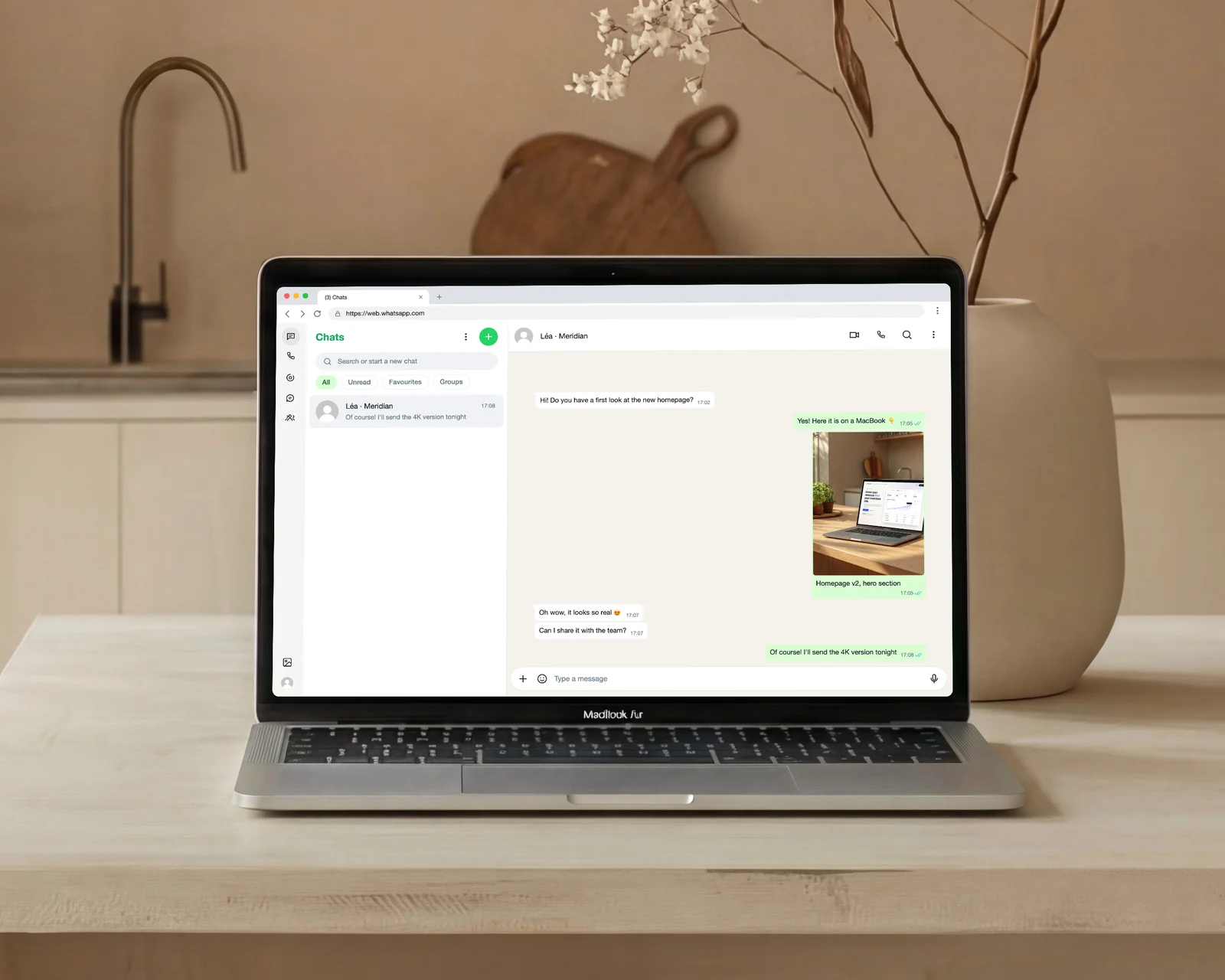 MacBook on a light wood counter in a minimalist kitchen showing the same conversation in WhatsApp Web