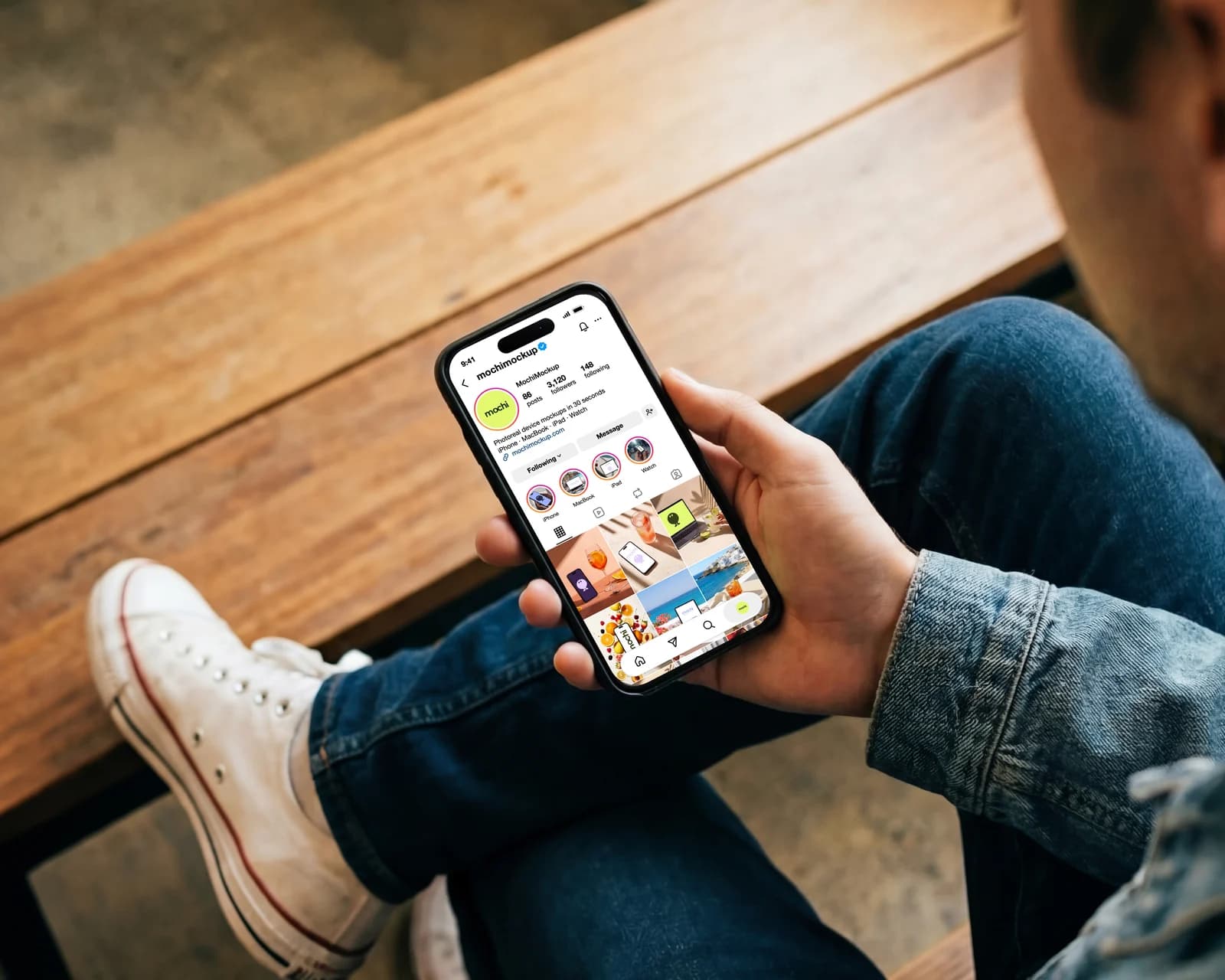 Man sitting on a bench holding an iPhone that shows the mochimockup Instagram profile with bio, highlights and photo grid